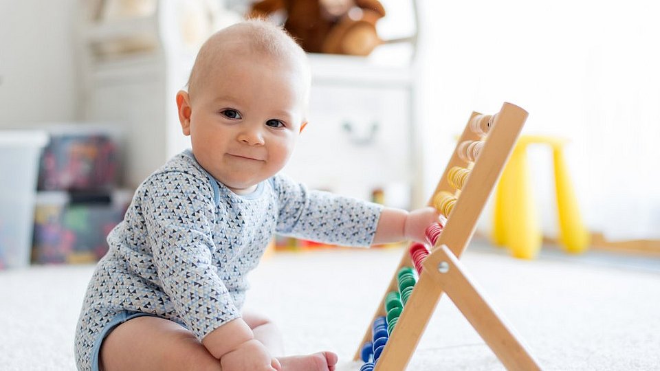 Baby spielt im Sitzen auf einem Teppich mit einem Holzspielzeug. 