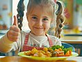 Ein Mädchen im Kindergartenalter sitzt am Tisch vor einem gefüllten Teller und zeigt die Daumen hoch-Geste mit einem Lächeln.