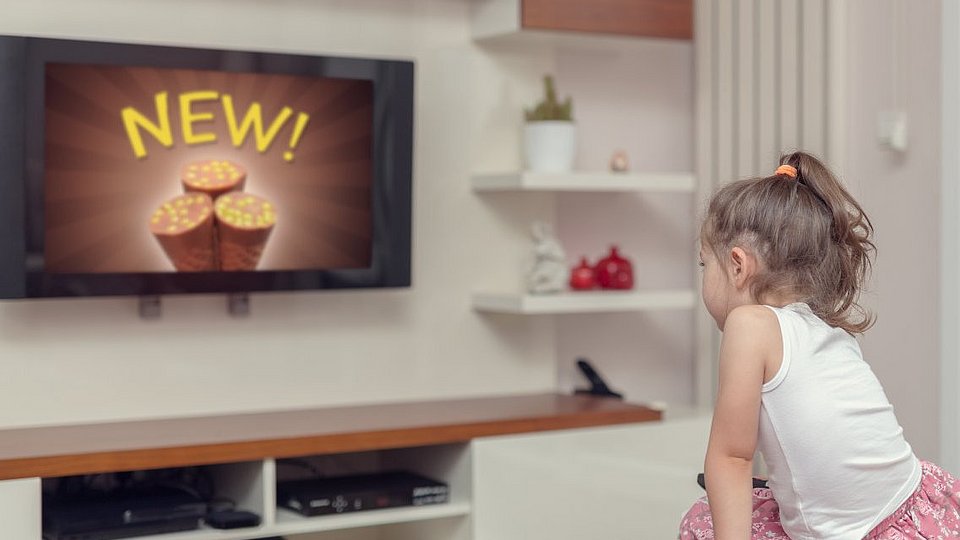 Kleines Mädchen sitzt vor dem Fernseher und schaut Werbung für Süßigkeiten. 