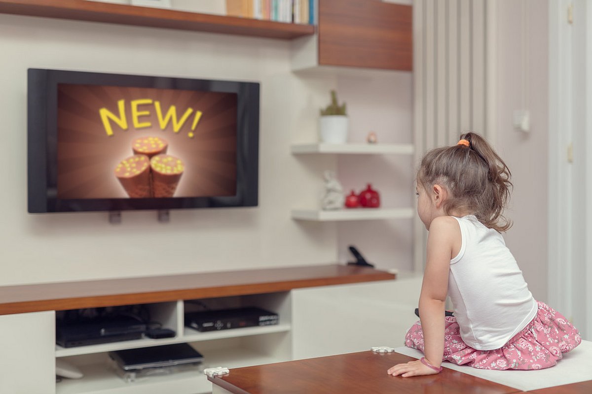 Kleines Mädchen sitzt vor dem Fernseher und schaut Werbung für Süßigkeiten. 