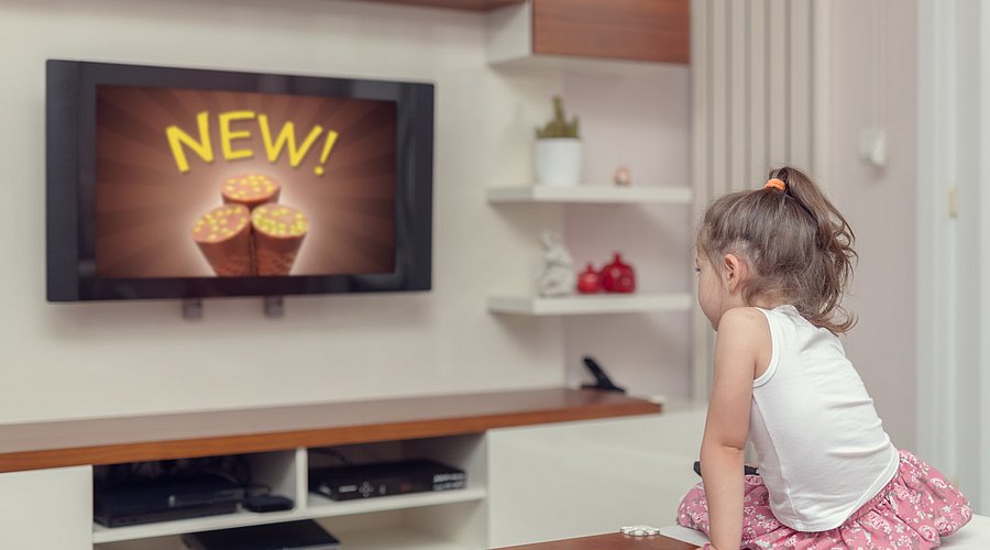 Kleines Mädchen sitzt vor dem Fernseher und schaut Werbung für Süßigkeiten. 