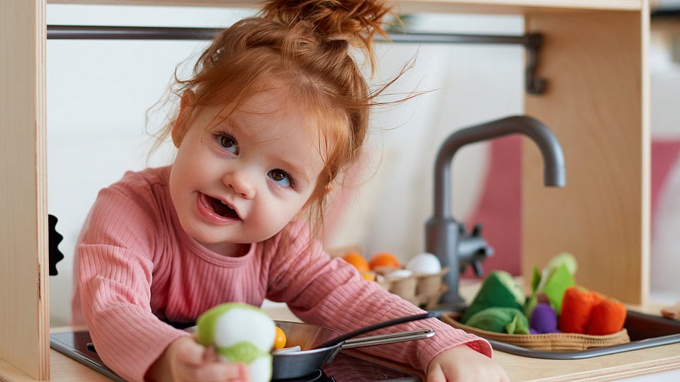 Ein Kleinkind spielt in einer Kinder-K&uuml;che mit Lebensmitteln aus Holz. 