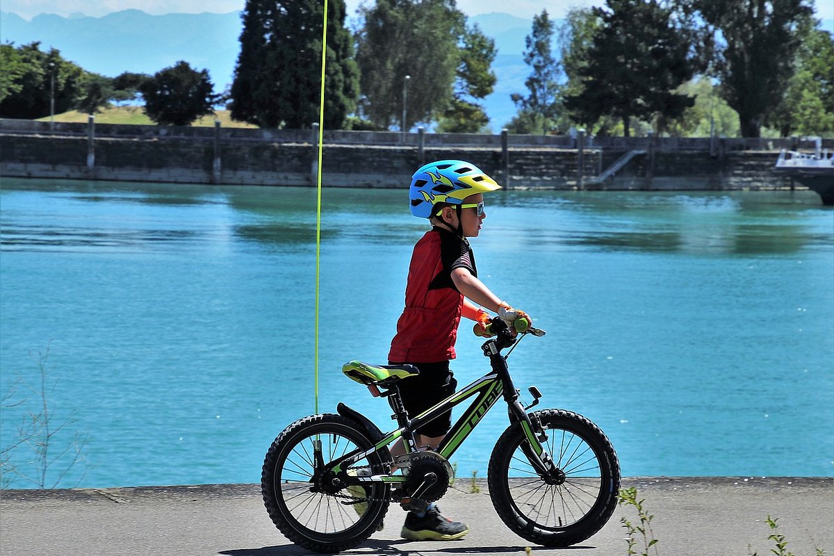 Ein Junge im Kitaalter steht neben seinem Fahrrad an einem See. Er trägt einen Helm, das Fahrrad hat einen Sicherheitswimpel.