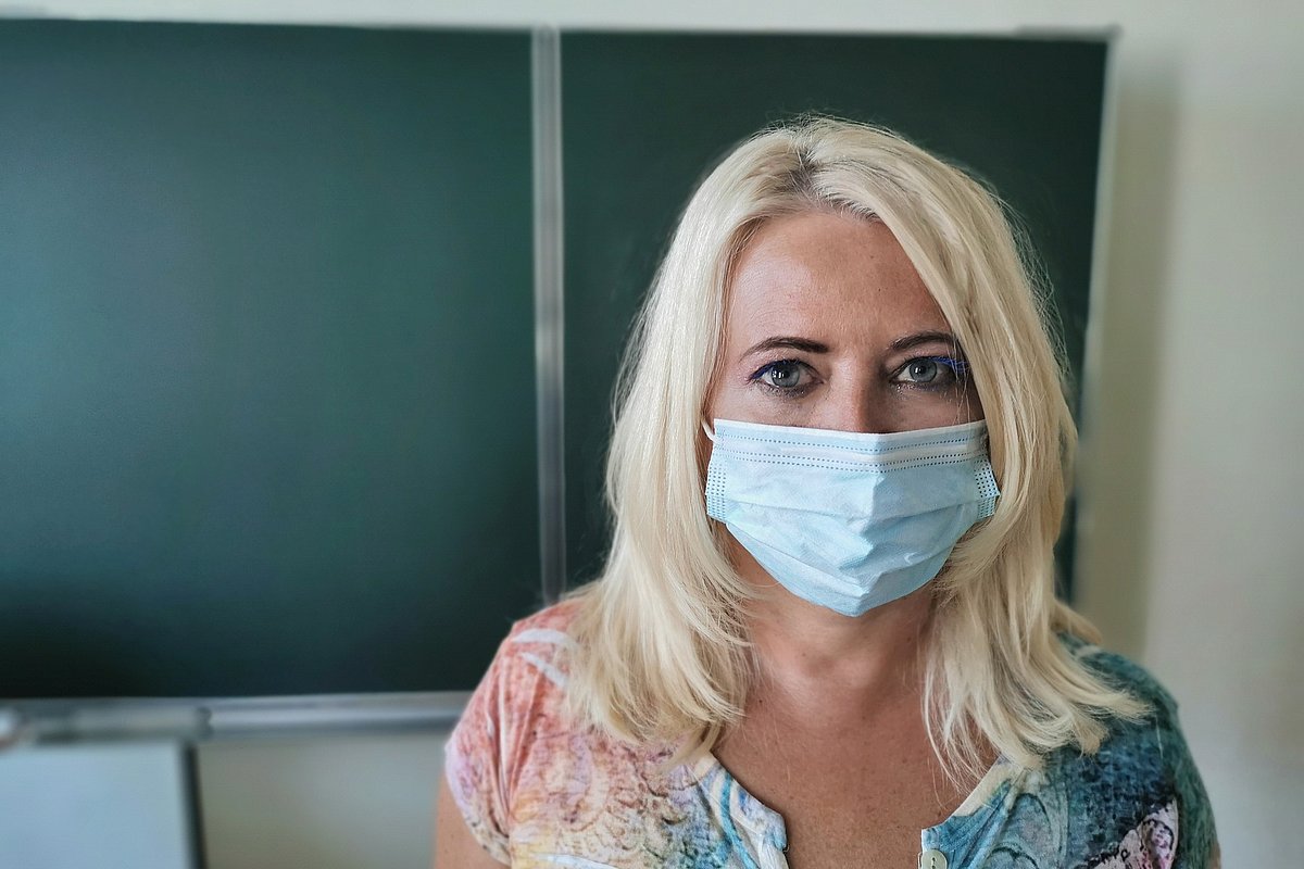 Eine junge Lehrerin mit Corona-Maske steht im Klassenraum vor einer leeren Tafel.