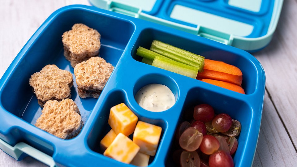 Eine geöffnete Brotdose zeigt Fächer mit gesunden Snacks für ein mitgebrachtes Mittagessen in einer Schule.