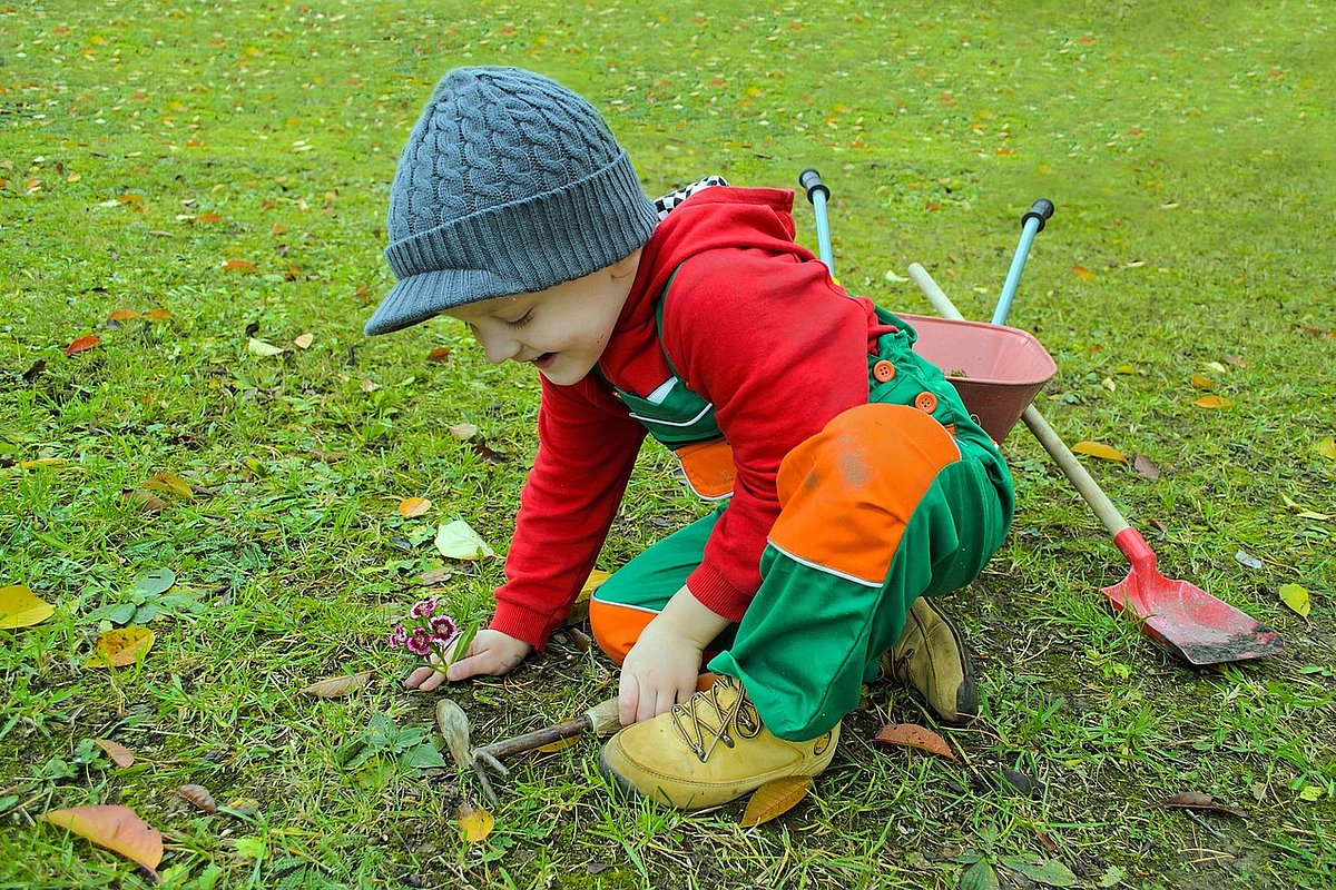 Ein Junge in Regenhose sitzt mit Gartenwerkzeug auf dem Rasen und pflanzt Blumen. 