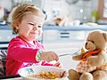 Kleines Mädchen sitzt im Hochstuhl am Tisch und füttert ihren Teddybären. 