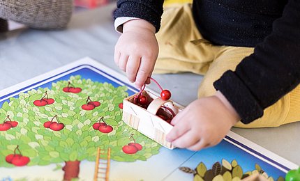 Ein Kleinkind spielt ein Brettspiel und packt Kirschen aus Holz in einen Korb.