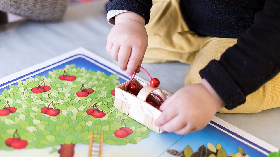 Ein Kleinkind spielt ein Brettspiel und packt Kirschen aus Holz in einen Korb.