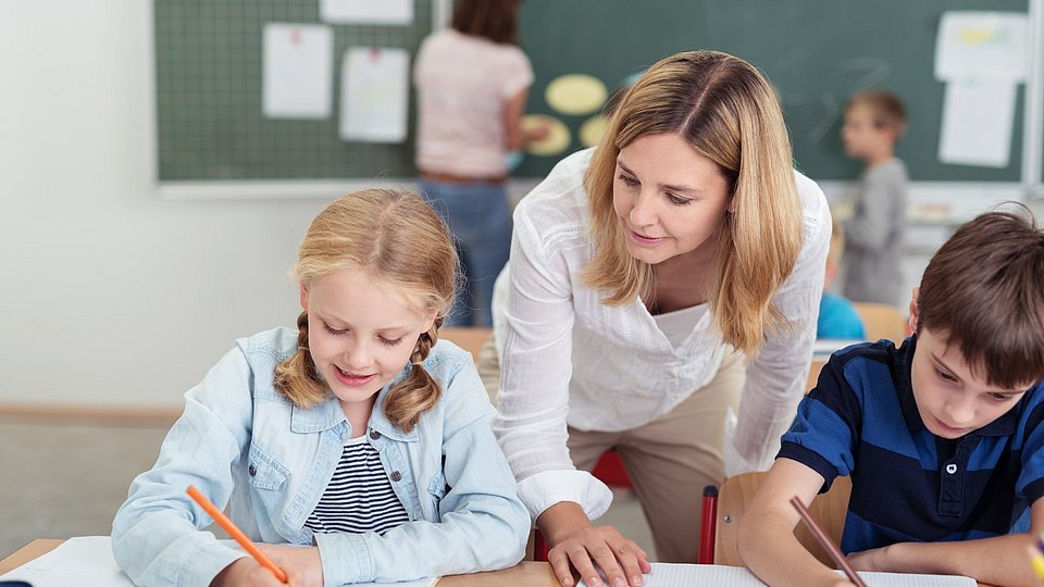 Die Szene zeigt eine Interaktion zwischen Lehrerin und zwei Grundschulkindern. 