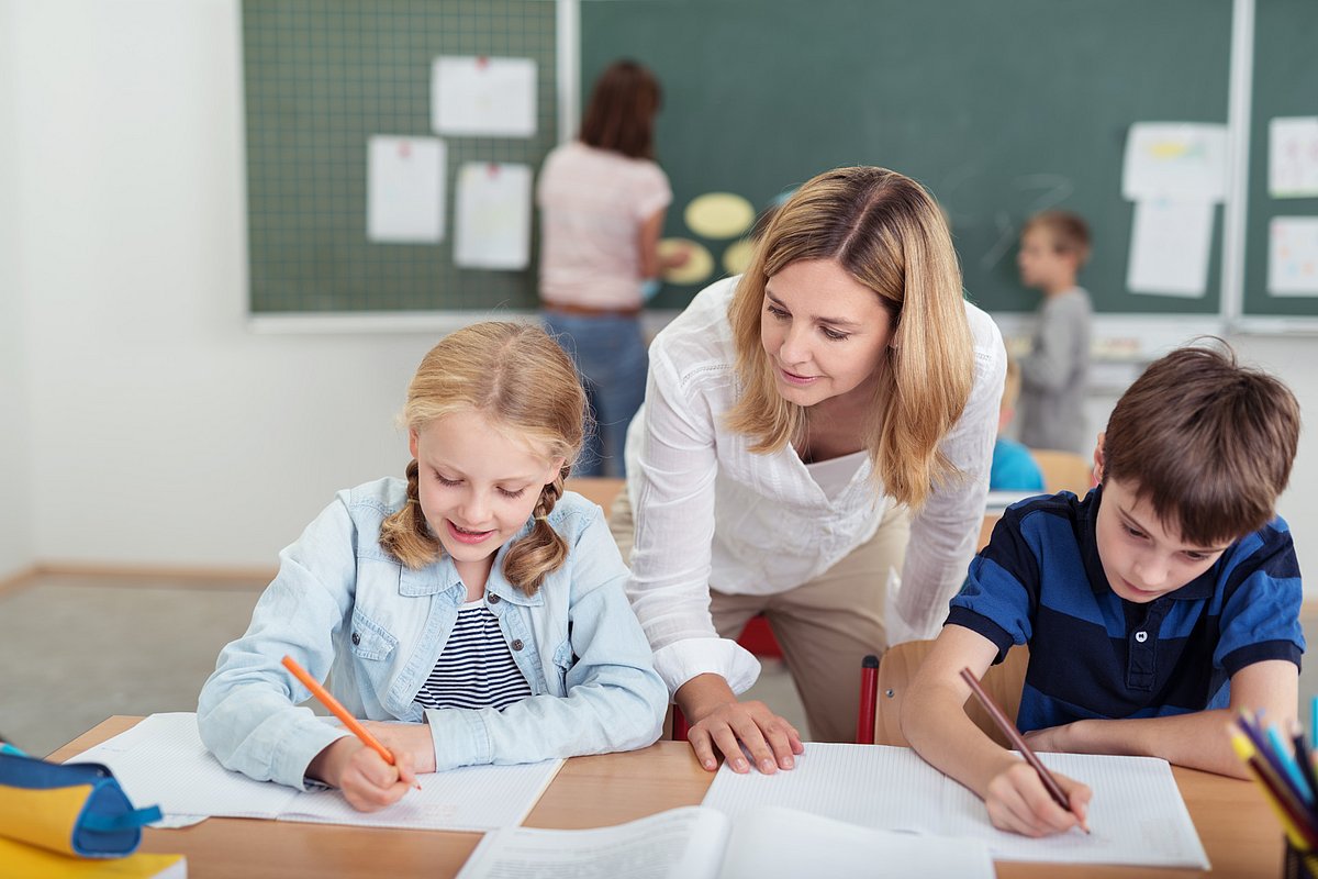 Die Szene zeigt eine Interaktion zwischen Lehrerin und zwei Grundschulkindern. 