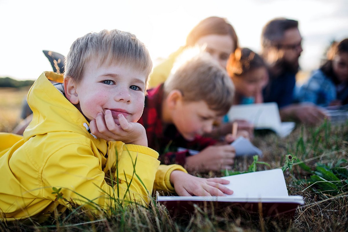 Kinder liegen mit ihrem Lehrer auf einer Wieser und haben Schulhefte vor sich.