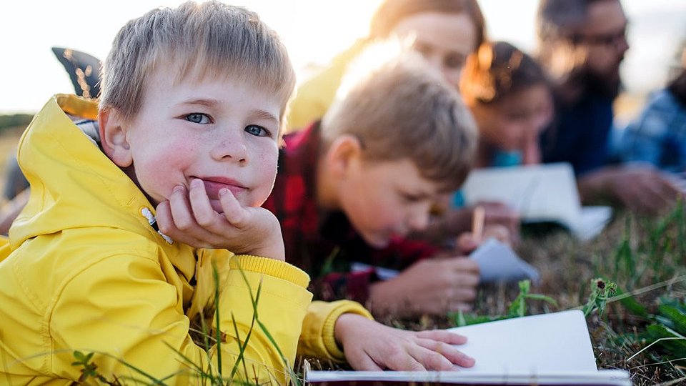 Kinder liegen mit ihrem Lehrer auf einer Wieser und haben Schulhefte vor sich.