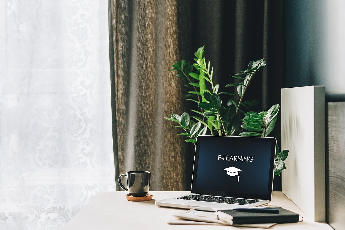 Arbeitsplatz mit einem aufgeklappte Laptop, auf dem Bildschirm steht E-Learning