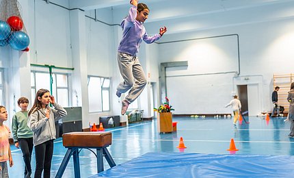 Schulkinder im Sportunterricht: Ein Mädchen springt von einem Sportgerät auf eine Weichbodenmatte. 