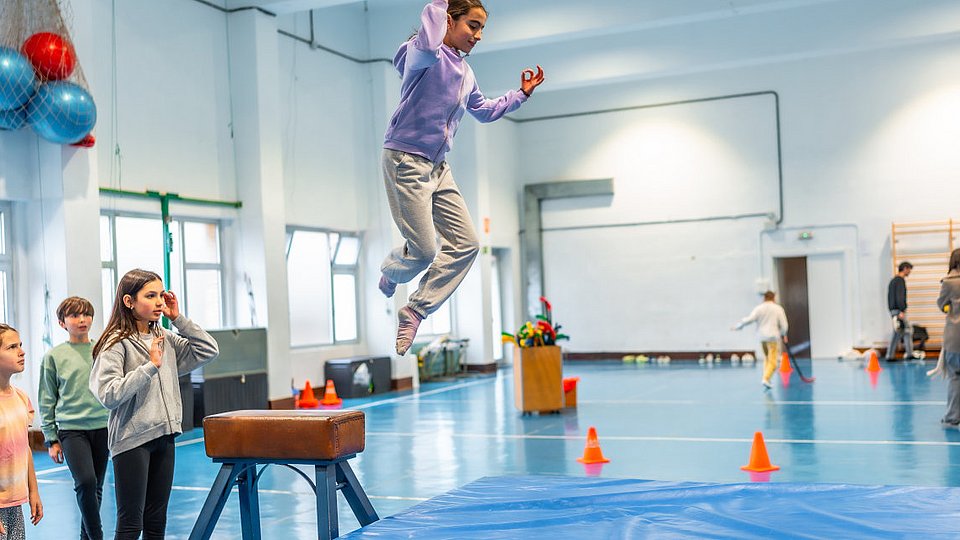 Schulkinder im Sportunterricht: Ein Mädchen springt von einem Sportgerät auf eine Weichbodenmatte. 