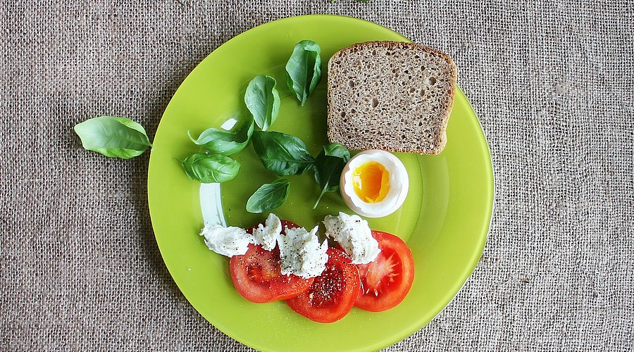 Auf einem grünen Teller sind drei Tomatenscheiben mit Feta-Käse, Basilikumblättern und einer Scheibe Vollkornbrot angerichtet. In der Mitte liegt ein halbes Ei. 