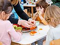 Kita-Kinder sitzen um einen Tisch und essen Obst von einer Obstplatte.