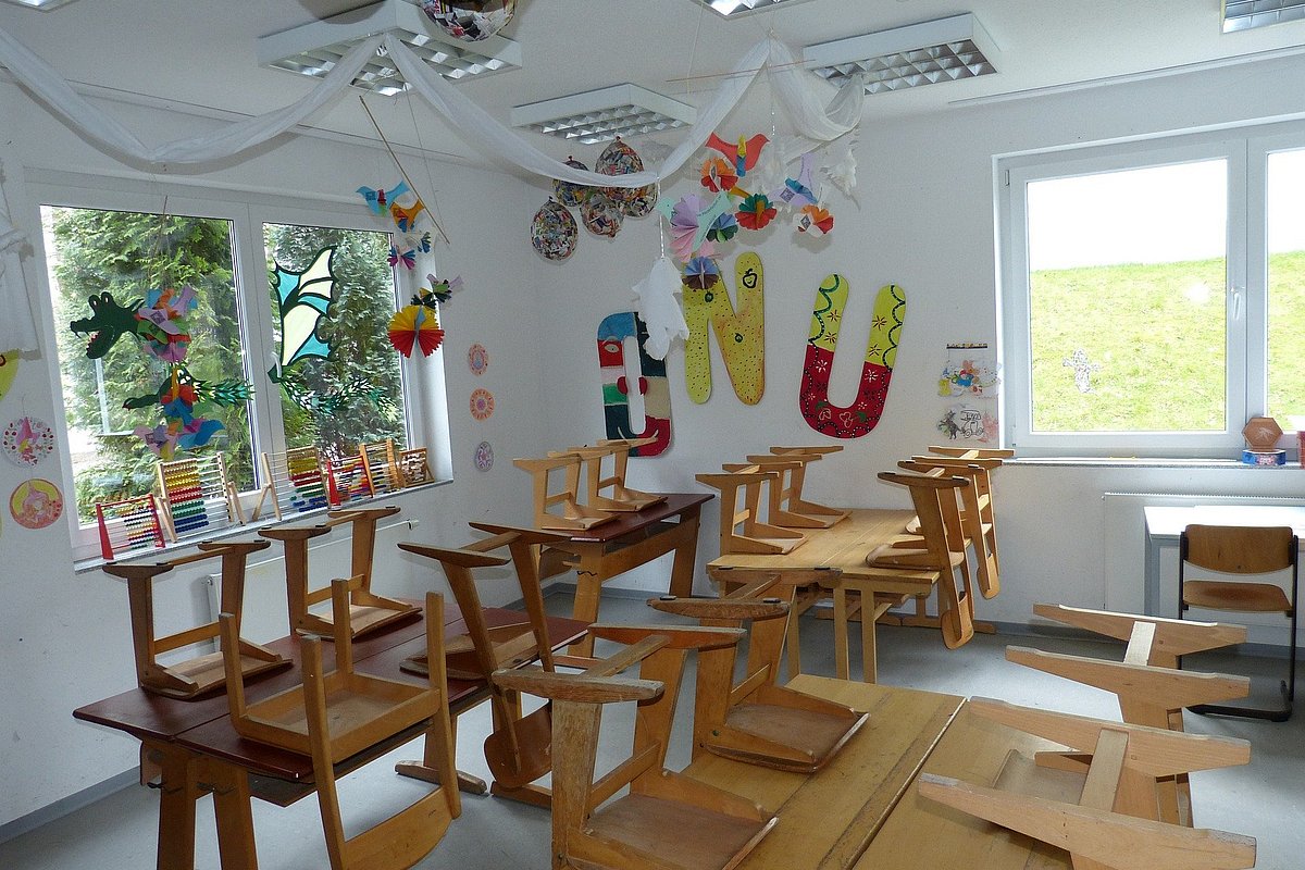 Ein leeres Klassenzimmer einer Grundschule mit, die Stühle sind hochgestellt. An den Wänden hängen bunte Bilder der Schülerinnen und Schüler.