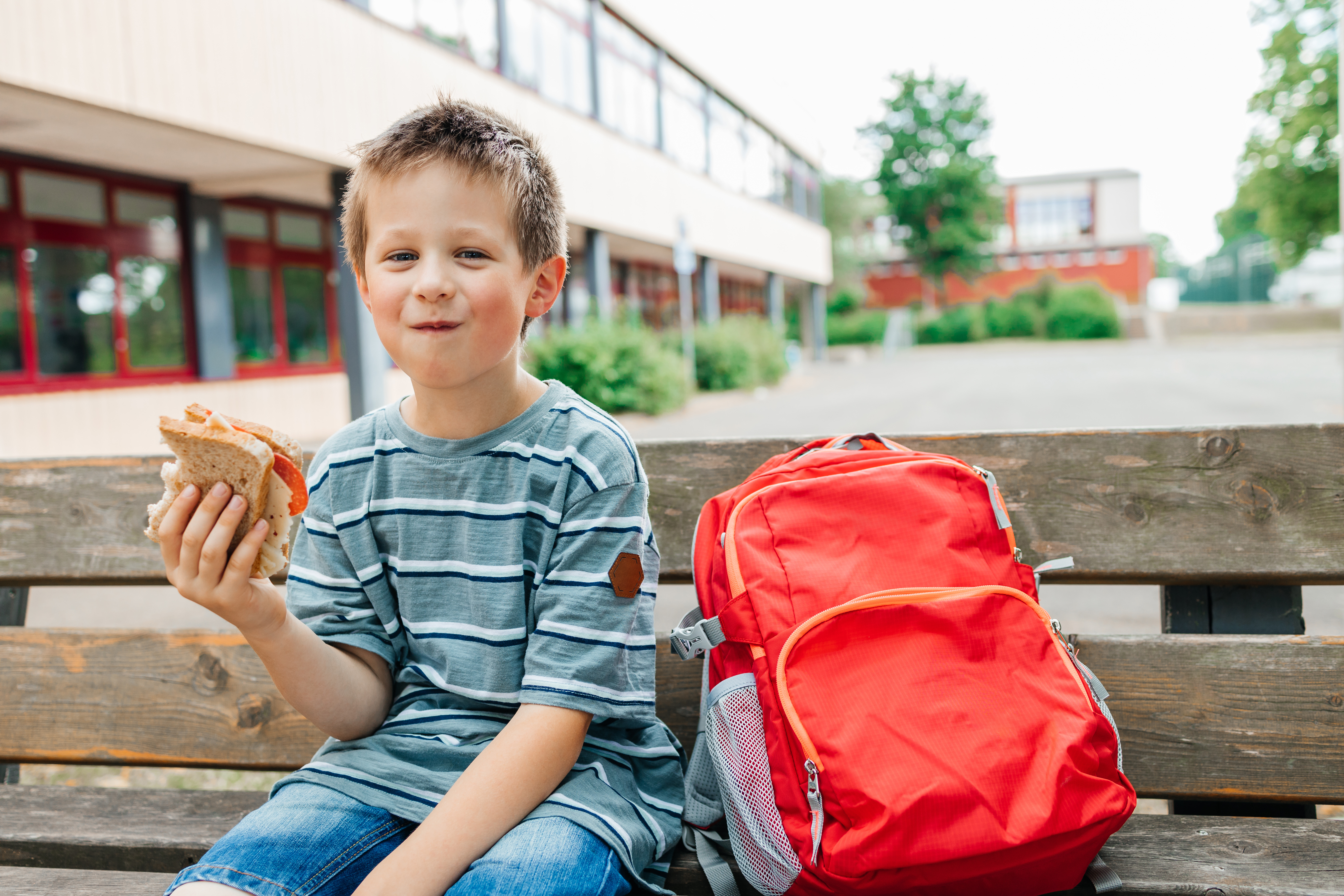 Ein Schuljunge mit kurzen blonden Stoppelhaaren sitzt verschmitzt lachend auf einer Holzbank vor seiner Schule und hält ein angebissenes Butterbrot in der Hand. Neben sich steht sein roter Schulrucksack.
